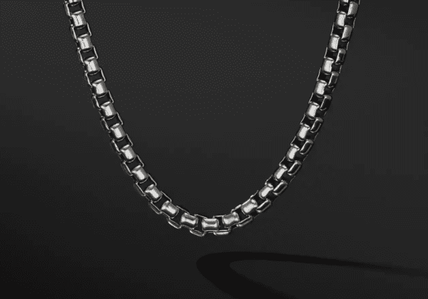 A close-up shot of the 5.2MM MENS BOX CHAIN displayed against a dark background. The chain showcases a box-link design, with each link forming small, interlocking squares that give it a sleek and polished appearance. The lighting accentuates the necklace's shine and texture beautifully.