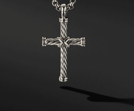 The CABLE CROSS pendant features a silver cross with an intricate, textured design and a woven pattern at the intersection of the two bars, hanging from a chunky chain. The dark background accentuates the detailed craftsmanship of the pendant.