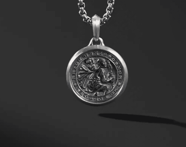 A close-up shot of the ST CHRISTOPHER pendant with intricate design, hanging from a chain. The pendant features an engraved image and detailed surrounding text. The background is dark, making the metallic pendant and chain stand out prominently.