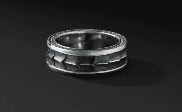 The 9MM ARMORY BAND, a silver ring featuring a textured, geometric design, is set against a black background. The ring boasts a wide band adorned with interlocking square and rectangular patterns around the middle, while its interior and edges have a smooth, polished finish.