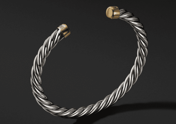 A close-up of the 6MM CABLE CLASSIC CUFF, showcasing its intricate braided design against a black background. The bracelet features gold-toned accents capping its ends.