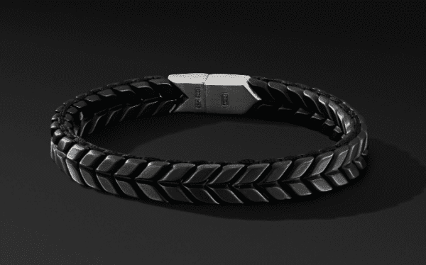 The 9MM Chevron Woven bracelet, featuring a black braided leather design and a silver clasp, is showcased against a dark background. Its interwoven pattern resembles herringbone or chevron styles, giving it a sleek and modern appearance.