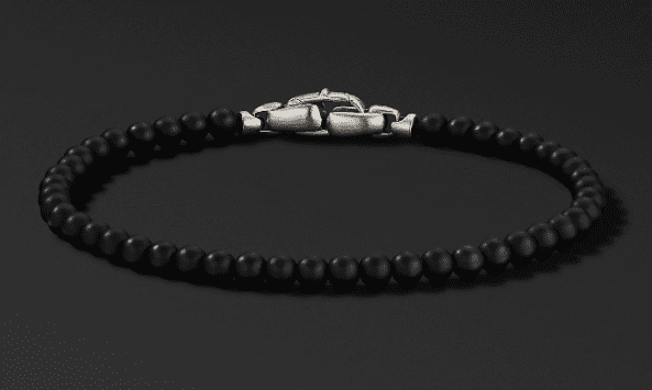 The 4MM SPIRITUAL BEAD bracelet features small, round black beads expertly strung together and fastened with a silver-toned clasp that resembles a chain link. Displayed against a dark background, this bracelet highlights its sleek and elegant design.