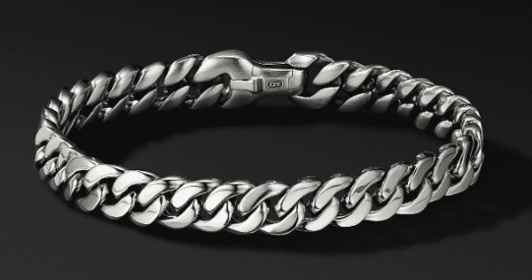 The 8MM CURB CHAIN BRACELET features a sophisticated design with interlocking chain links arranged in a circular form, set against a dark background. The clasp is intricately crafted and includes a small engraved detail, adding a touch of elegance to its sleek silver appearance.