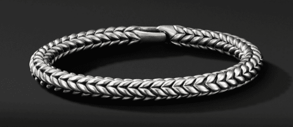 The CHEVRON BRACELET, featuring an intricate, braided design, is displayed against a dark background. The bracelet is closed by a secure, matching clasp that highlights its craftsmanship and elegant appearance.