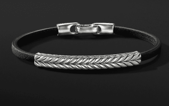 The CHEVRON LEATHER BRACELET is a sleek black leather accessory with a silver clasp, featuring a central silver accent with a braided design set against the dark leather.