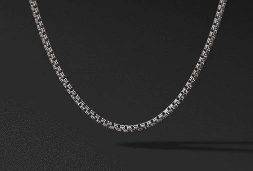 A 2.6MM DOUBLE BOX CHAIN necklace is displayed against a dark background. The chain features an intricate, closely linked design that creates a subtle texture. The lighting highlights the shiny, metallic surface, emphasizing the craftsmanship and elegance of the chain.