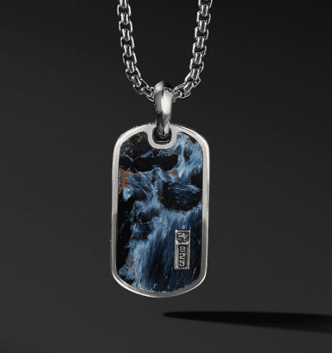 The EXOTIC STONE TAG, a silver rectangular pendant with a chain necklace, showcases a dark iridescent surface that shimmers with hints of blue and red. The pendant is adorned with a small rectangular silver tag featuring the engraving "FRED." This elegant piece is presented against a dark, minimalist background.