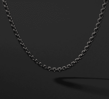 A close-up photo of the 2.7MM MENS BOX CHAIN necklace against a dark background. The silver chain forms a smooth, symmetrical curve, and the links are clearly visible, reflecting light subtly. The plain black background features a soft shadow cast by the chain, highlighting its elegance.