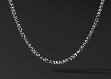 The 2.6MM DOUBLE BOX CHAIN, crafted from interlinked rectangular-shaped links, is displayed against a black background. The necklace forms a gentle curve, showcasing its intricate design and metallic luster.