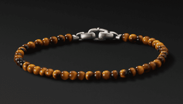 The 4MM SPIRITUAL BEAD bracelet, featuring polished brown stones interspersed with a few darker beads and held together by a chunky metallic clasp, is displayed against a dark background.