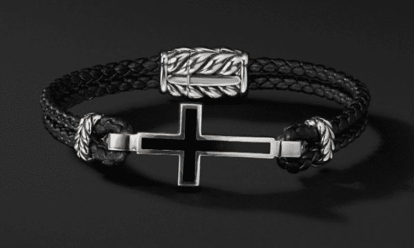 The CROSS STATION MED BRACELET is a black braided leather bracelet featuring a silver cross charm with black inlay. It also has silver accents with engraved patterns near the clasp, set against a solid black background.