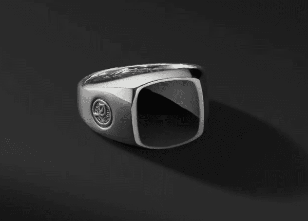 The 12MM CUSHION SIGNET RING BO showcases a striking silver band with a bold, square-shaped black gemstone set prominently in the center. The ring is enhanced by a delicate, engraved circular design on one side of the thick band, all set against a dark gradient background.