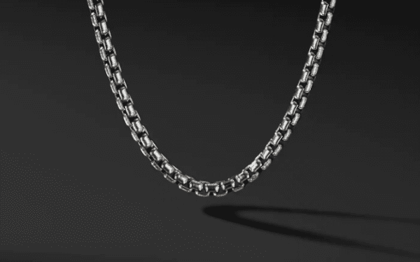 A close-up of the 4MM MENS BOX CHAIN set against a black background. The interlocking links create a repeating pattern that casts a subtle shadow, emphasizing the texture and shine of the metal. The focus is on the chain's craftsmanship and detail.