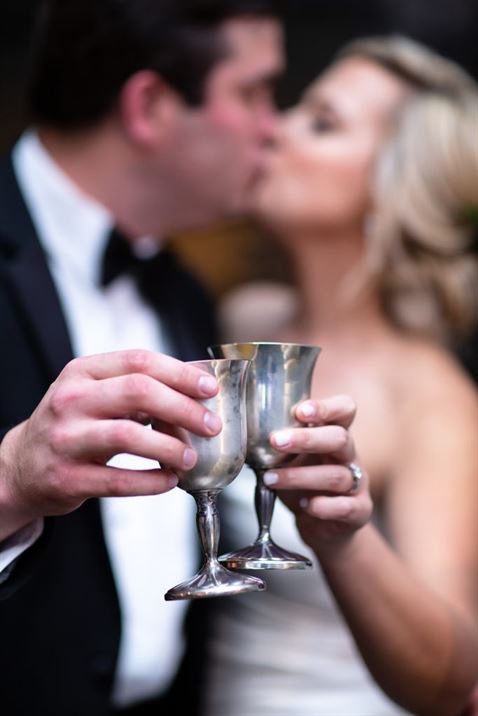 A Bromberg’s bride, wearing a white strapless wedding gown and diamond ring, and groom, wearing a tux, cheers with silver goblets.