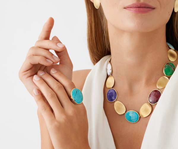 A woman adorned with a gold necklace featuring vibrant gemstone pendants and the Marco Bicego Lunaria Turquoise Cocktail Ring elegantly completes her look. She has long hair, a sleeveless white top, and gracefully positions her hands near her face.