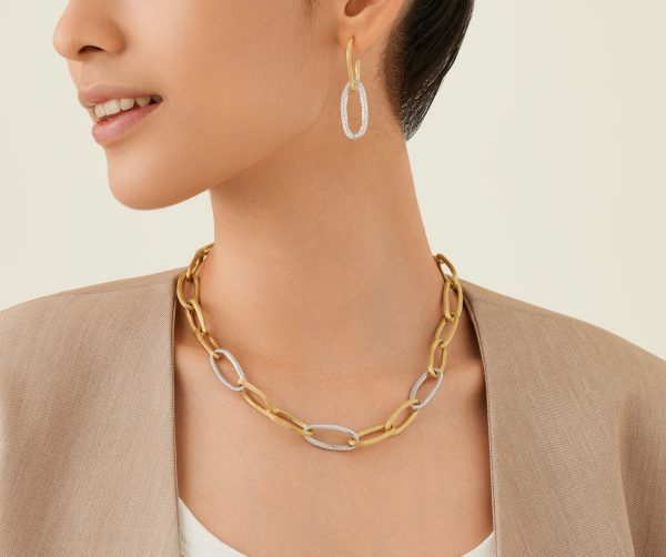 A person wearing gold and silver oval-shaped earrings along with the Marco Bicego Jaipur Diamond Link Necklace, featuring alternating large links. They are dressed in a beige blazer over a white top, against a light beige background.