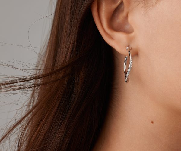 Close-up of a person wearing the elegant Marco Bicego Marrakech Diamond Twist Hoop Earrings in sleek silver. Their dark brown hair partially covers their ear, set against a neutral background.