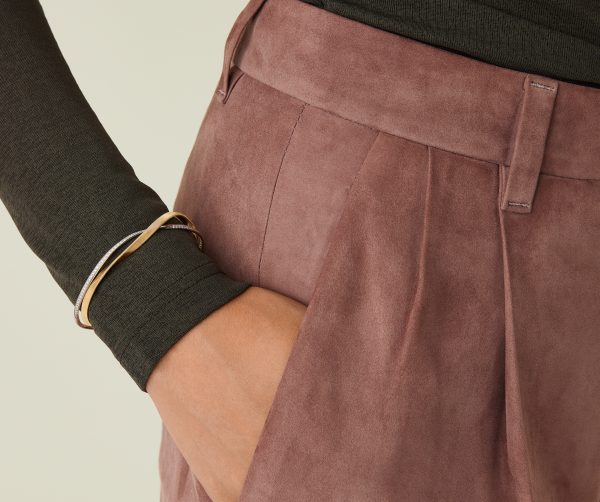 Close-up of a person wearing a dark long-sleeve top and dusty pink suede trousers, with one hand in the pocket. They are adorned with a Marco Bicego Marrakech Two-Strand Diamond Bangle on their wrist. The textures of the clothing are clearly visible.