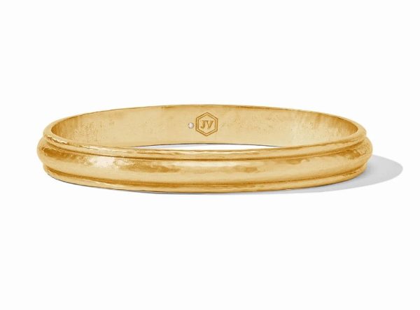 The Julie Vos Cirque Bangle Bracelet features a gold finish with a hammered texture and a small emblem showcasing the initials "JV" engraved inside. The bracelet has a slightly reflective surface and boasts a simple, elegant design. It is displayed on a white background, casting a subtle shadow.