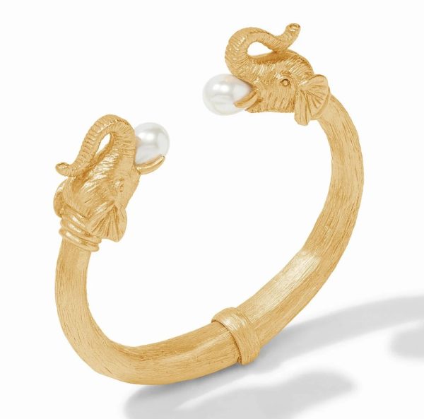 The Julie Vos Elephant Cuff is a gold bangle bracelet that showcases two elephant heads facing each other at the open ends, each gracefully holding a pearl in its trunk. The bracelet features a textured finish and casts a striking shadow on the white background.