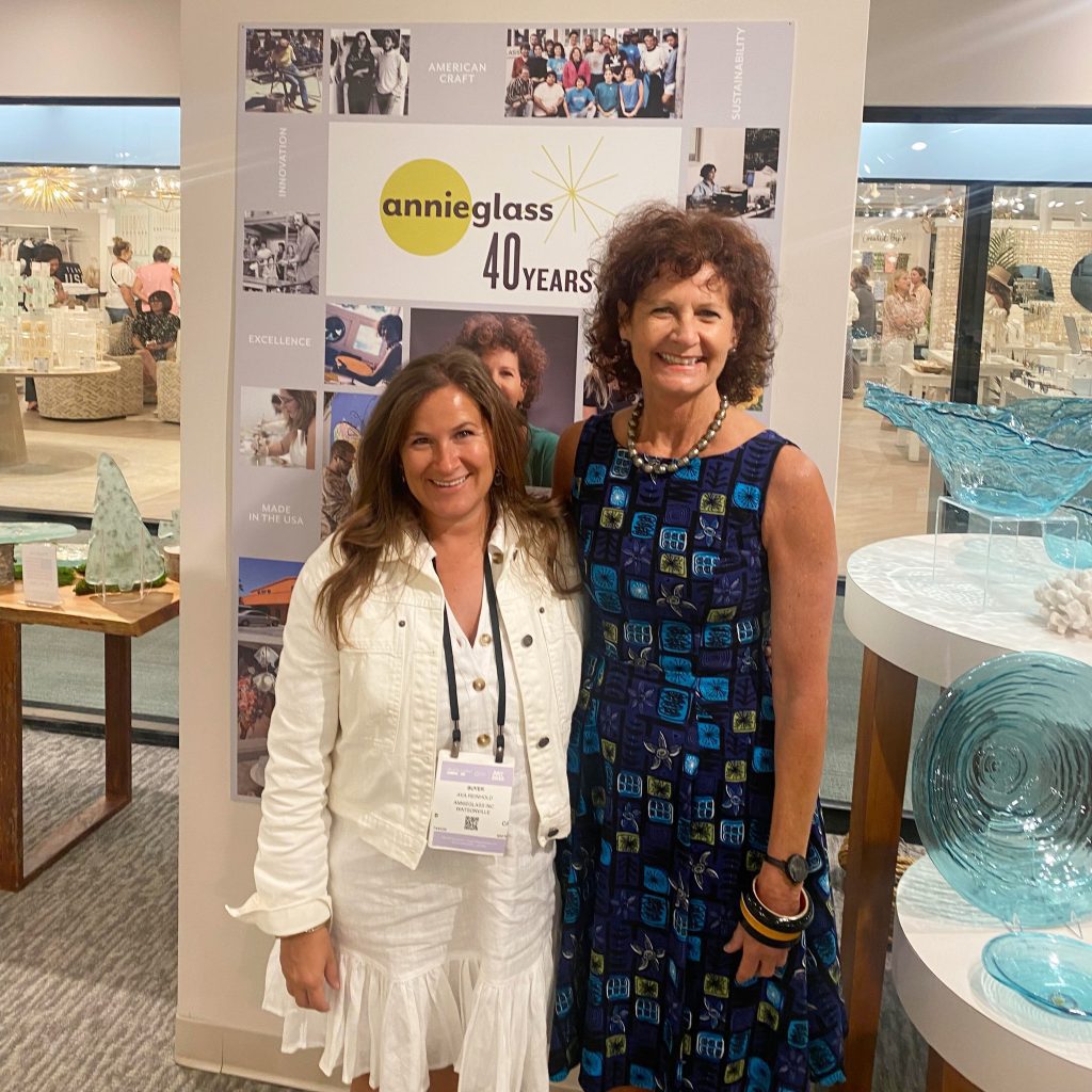 Two women smile while standing in front of a display celebrating the 40th anniversary of Annieglass. The display features various images and the Annieglass logo. One woman is wearing a white dress and jacket, and the other is in a patterned blue dress.