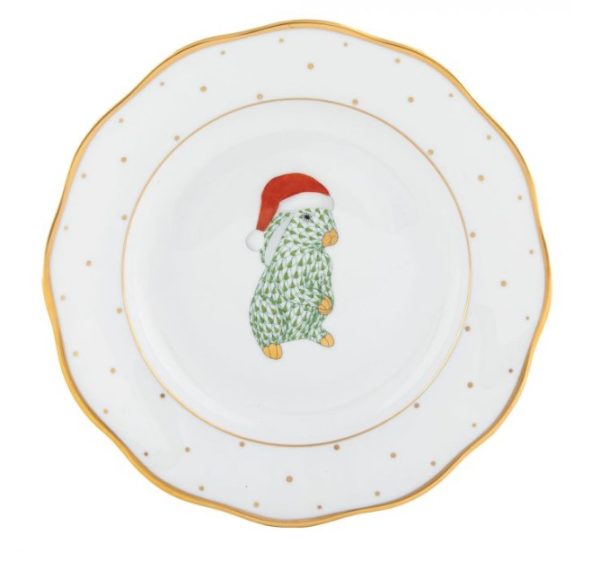 The Herend Christmas Dessert Plate - Santa Bunny is a white plate with a scalloped edge trimmed in gold. It features a small illustration of a green cactus wearing a red and white Santa hat in the center, with the cactus arranged in a decorative pattern resembling a Christmas tree. Additionally, small gold dots are scattered around the rim.