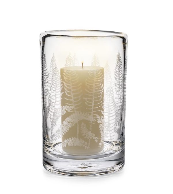 The Simon Pearce Fern Hurricane features a lit white candle enclosed in a clear glass holder adorned with an etched fern leaf design, casting a warm glow that highlights the intricate fern patterns on the glass.