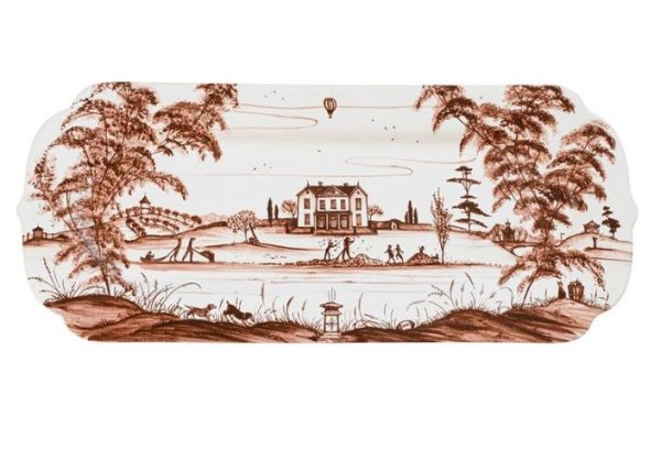 The Juliska Country Estate Harvest 15" Hostess Tray in Sepia features an elongated porcelain design adorned with a detailed landscape painting. The scene showcases a grand house encircled by tall trees and a lush garden. In the background, a hot air balloon drifts through the sky, while in the foreground, there is a small lake complete with a boat and lighthouse.