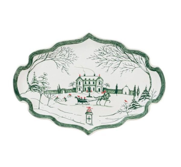 The Juliska Country Estate Winter Frolic 7" Tray in Evergreen is an ornate oval-shaped ceramic platter that features a winter scene of a mansion surrounded by snow-covered trees. In the foreground, horse-drawn sleighs add to the image's festive, holiday feel.