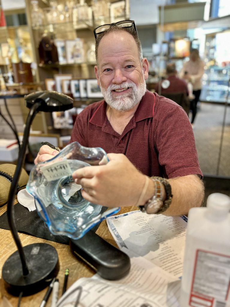 Master Engraver at Bromberg’s in Birmingham, AL, smiles while engraving a crystal vase at his workstation.
