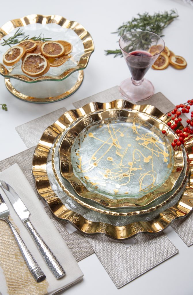 A dining table set with elegant golden-rimmed plates, a glass of red wine, and cutlery on a beige placemat. A glass dish with dried citrus slices and a small branch with red berries complete the festive look.