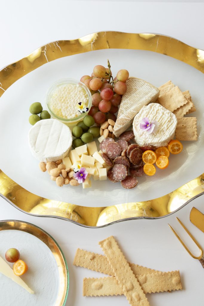 A decorative platter with various cheeses, green olives, grapes, nuts, salami slices, and crackers. An elegantly placed small glass bowl contains a spread topped with a flower. Nearby, a small dish holds a single cracker and garnish.