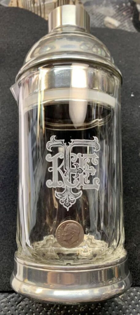 A shiny, clear glass stein with custom-engraved decorative monogram letters "RCE" on its body. The silver-toned lid is closed, and there's a coin placed at the base inside the stein to provide a sense of scale. The stein stands on a textured black surface.