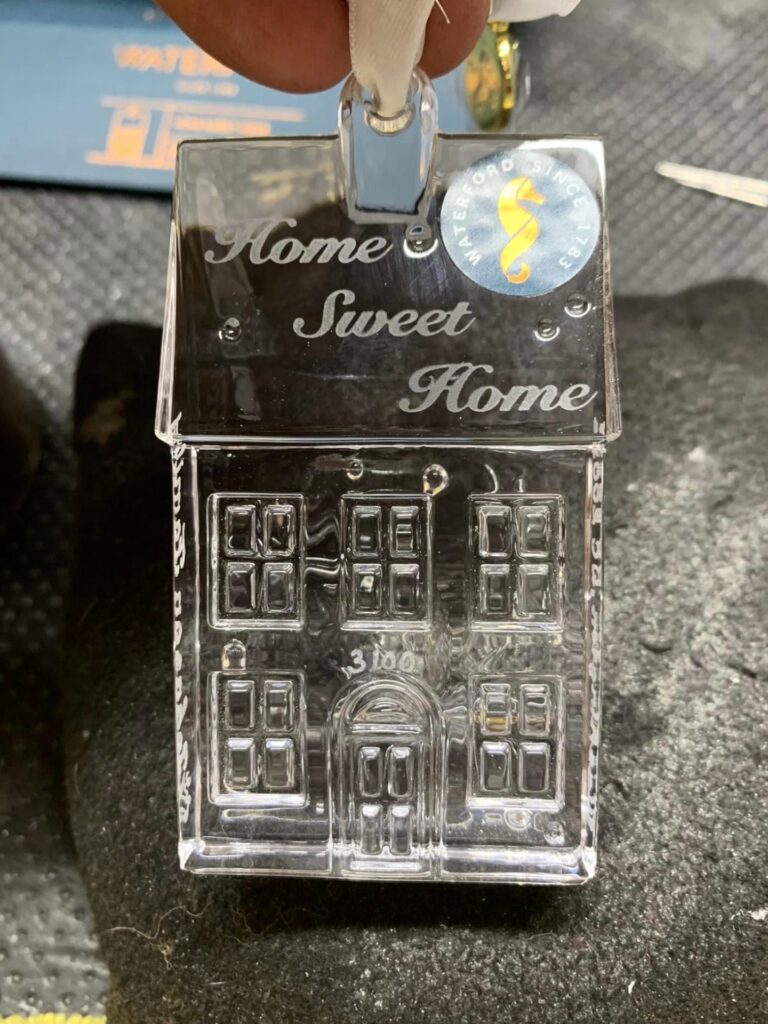 A clear, detailed glass ornament in the shape of a house is held up by a hand. The house features intricate details like windows, a door, and an overhanging roof with custom engraving "Home Sweet Home." A round blue sticker with a yellow symbol adds to its charm.