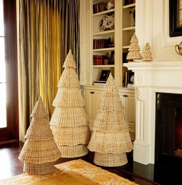 A cozy living room with Juliska Provence Rattan Trees - 5 ft in various sizes placed near a fireplace. Shelves filled with books and decorative items are in the background, alongside a large window with heavy drapes. A small Juliska Provence Rattan Tree is on the fireplace mantel.