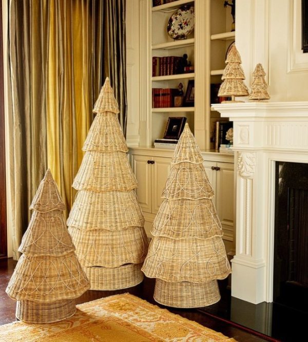 A living room with a Juliska Provence Rattan Tree - 3 ft, along with two other wicker Christmas tree decorations of varying sizes, placed near a fireplace. The fireplace mantel features smaller wicker tree decorations and a white, ornately carved design. A curtain to one side covers a window, and a rug adorns the floor.