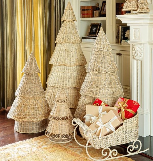 Four tiered wicker Christmas trees in varying sizes are displayed in a cozy living room. Beside them, the Juliska Provence Rattan Sleigh filled with wrapped gifts is positioned. In the background are a fireplace, bookshelves, and curtains.