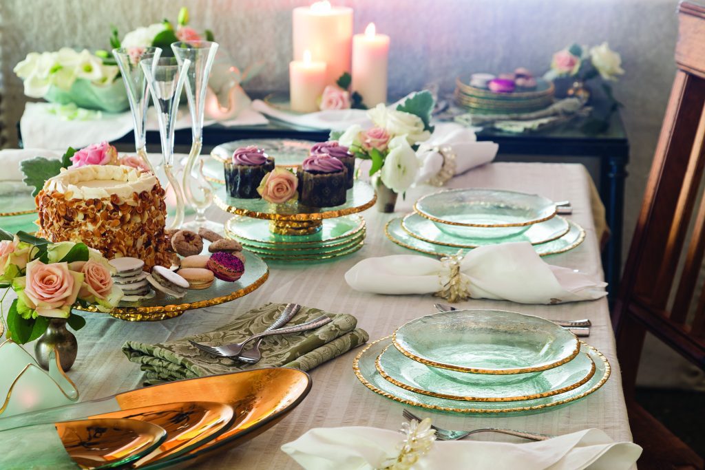 A beautifully set dining table with green and gold rimmed plates, folded cloth napkins, and silver cutlery. The table features a cake, cupcakes, macarons, floral arrangements, and lit candles in the background, creating a cozy and elegant ambiance.