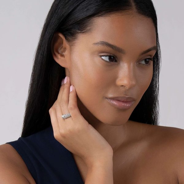 A woman with long, smooth hair and a serene expression gently touches her neck, showcasing the LAGOS Embrace X Beaded Ring on her finger. She is wearing a dark, sleeveless top and has a soft, natural makeup look with a subtle sheen on her skin. The background is plain and light.