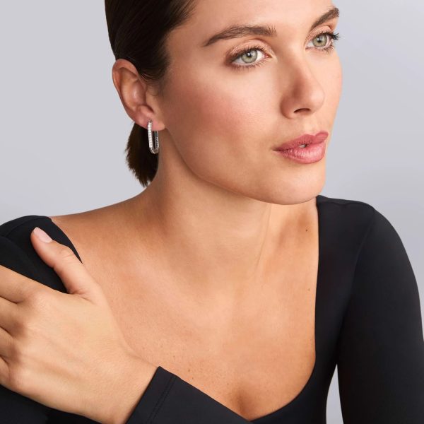 Woman with light skin and green eyes wearing a black top and LAGOS Caviar Spark Linear Diamond Hoop Earrings, looks to the side and holds one arm across her chest. The background is a soft gray.