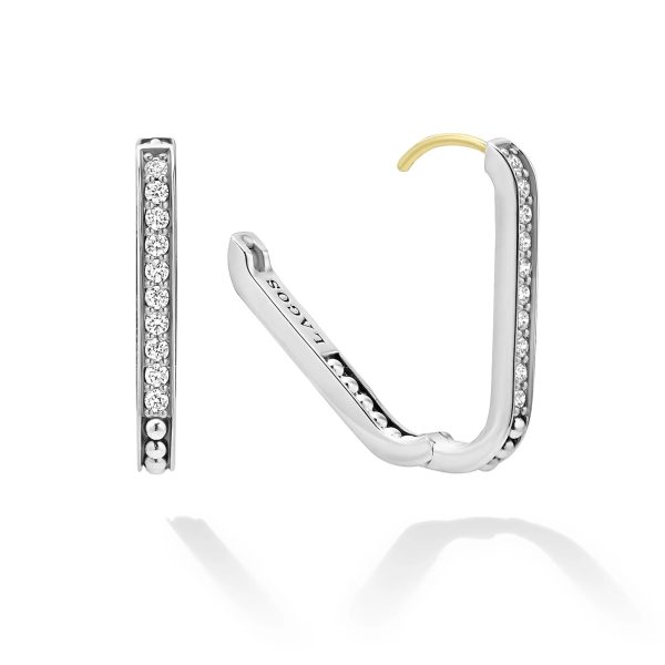 Displayed is a pair of LAGOS Caviar Spark Linear Diamond Hoop Earrings. They are silver with a row of small diamonds set along the outer rim. The open earring reveals a clasp mechanism with a touch of gold at the hinge. The brand name "Lagoss" is engraved inside. The reflection of the earrings can be seen on the surface below them.