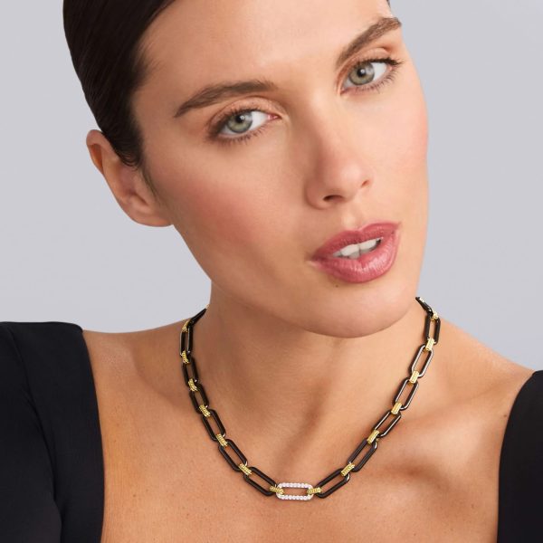 A woman with light skin and green eyes is wearing a black top and showcasing the LAGOS Signature Caviar 18K Gold and Black Ceramic Diamond Station Link Necklace. She has her hair pulled back and is set against a plain, light-colored background.