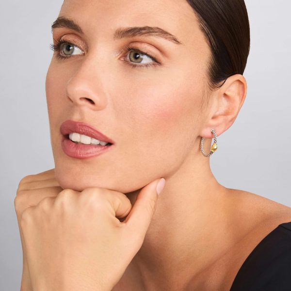 A person with fair skin and dark hair pulled back is wearing LAGOS Signature Caviar Two-Tone Station Hoop Earrings, which feature a delicate twisted design with a small gold-colored charm. They are resting their chin on their hand and gazing slightly upward with a serene expression.