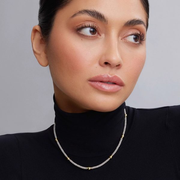 A woman with tied-back dark hair looks to her left, wearing a black turtleneck and a delicate LAGOS Signature Caviar 18K Gold Station Necklace. The close-up showcases her minimal makeup and the elegant simplicity of her attire and accessories.