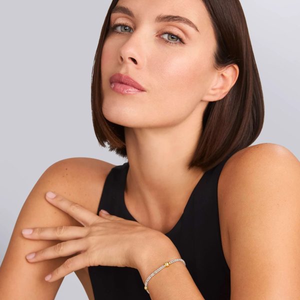 A woman with short, dark hair wearing a sleeveless black top gazes confidently at the camera. She rests her right hand on her shoulder, subtly showcasing the Signature Caviar Two-Tone Station Bracelet on her wrist. The background is a neutral gray.