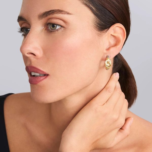 A person with fair skin and dark hair tied back is shown in profile, wearing LAGOS Signature Caviar Two-Tone Interlocking Diamond Hoop Earrings. The person is touching their neck with their hand and looking ahead. They are dressed in a black outfit and have a neutral facial expression.