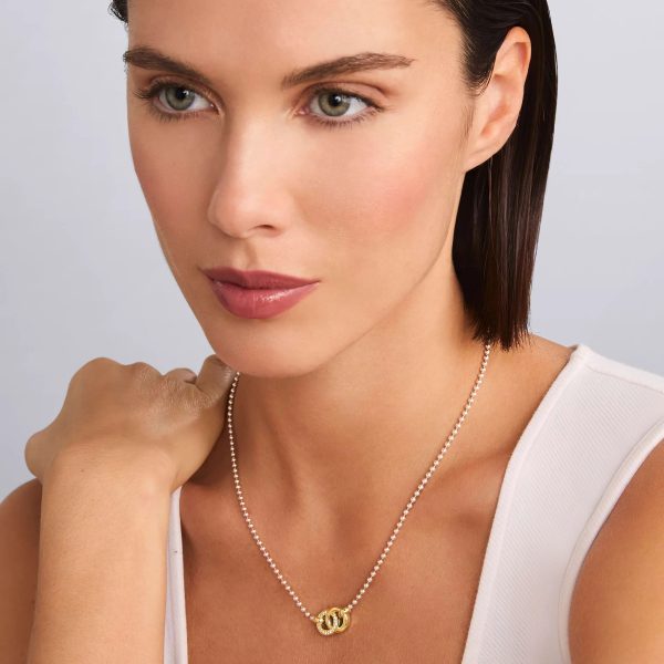 A woman with short brown hair wears a white sleeveless top and the LAGOS Signature Caviar Two-Tone Interlocking Diamond Pendant Necklace. She gazes slightly to the side with a calm expression and has her hand resting near her chin against a neutral background.