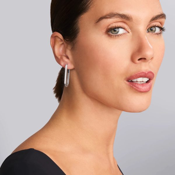 A woman with light skin and green eyes poses against a gray background. She wears LAGOS Caviar Spark Large Linear Diamond Hoop Earrings and a black top, with her brown hair pulled back. She gazes toward the camera with a neutral expression.