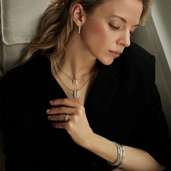 A woman with long, blonde hair is sitting in a relaxed pose, looking down. She is wearing a black top and accessorized with silver jewelry, including hoop earrings, two layered necklaces—one being the LAGOS Caviar Spark Large Linear Diamond Pendant Necklace—bracelets, and a ring. The background is softly lit and neutral in color.